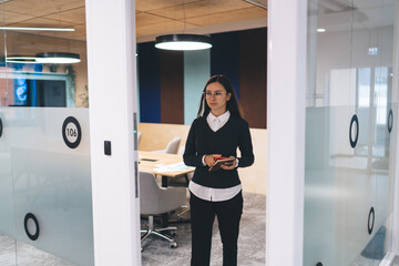 Young Hispanic woman in smart casual outfit enters modern office meeting room holding notebook,...
