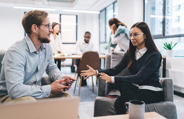 Focused young man explaining ideas to a woman in a lounge area of a coworking office, emphasizing leadership, business consulting, and strategic planning with digital communication tools.
