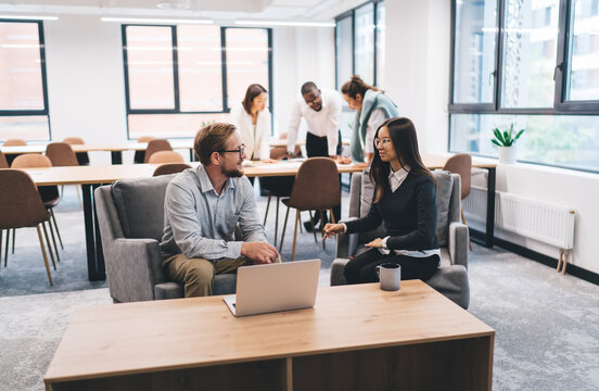 Caucasian man and Asian woman seated in open-plan office lounge discussing project strategy, with laptop and coffee mug on table, portraying teamwork and digital-era leadership exchange
