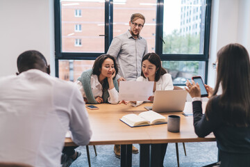 Two female professionals attentively reviewing printed report while male colleague stands behind them, actively participating in data analysis during collaborative office session