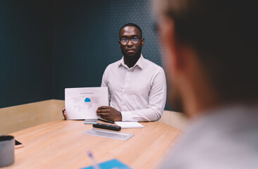 Serious African-American businessman presenting printed chart in boardroom, sharing cloud computing...