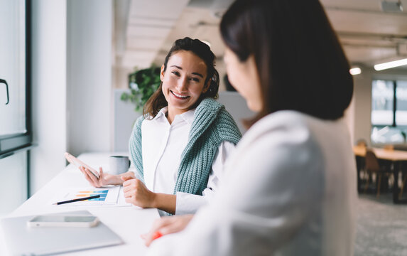 Smiling young woman having friendly conversation with colleague in bright modern office. Casual business atmosphere, digital chart and smartphone on desk