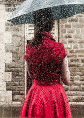 A woman in a red dress stands in the rain under an umbrella.