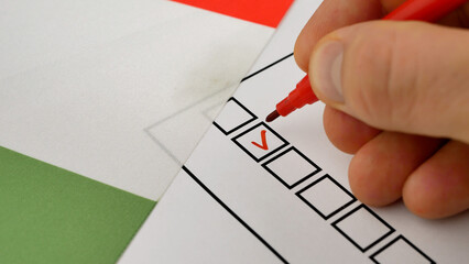 Voter places red marker check beside candidate name. Elections in Italy. National flag of Italian Republic visible in background. Secure voting process ensures fair political representation