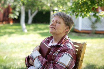 Senior woman in a checkered jacket enjoying a peaceful sunny day with closed eyes and holding a book to her chest, concept of relaxation and mindfulness in old age