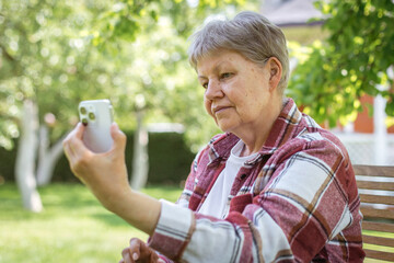 Senior woman in plaid shirt and white t-shirt sitting outdoors on sunny day, holding smartphone and making video call, concept of communication, theme of technology and aging