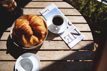 Sunlight-lit breakfast setting with flaky croissants, black coffee, and a newspaper on a wooden...