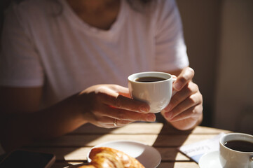 Close-up of hands holding a cup of coffee with a croissant on the table. A cozy moment enjoying breakfast in a bright and warm setting.