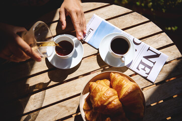Glasses and cups of coffee with fresh croissants on a wooden outdoor table, bathed in soft sunlight, evoke a feeling of a relaxed morning or social cafe atmosphere.