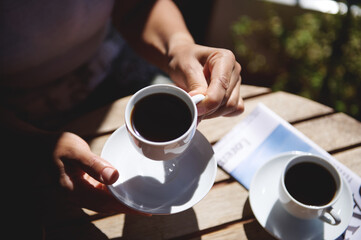 A person savoring a cup of coffee outdoors at a sunlit table, accompanied by a magazine and another cup. The peaceful ambiance underscores leisurely mornings and outdoor relaxation.
