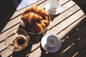 Warm golden croissants served in a bowl, accompanied by coffee cups, capture the cozy morning mood, set on a rustic wooden table bathed in soft sunlight.
