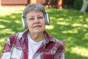 Senior woman in red plaid shirt and white t-shirt sitting on chair in garden with headphones, enjoying calm moment on sunny day, concept of relaxation, theme of aging, leisure, lifestyle