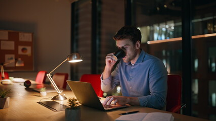 Overtime manager drinking coffee looking laptop screen at dark office. Tired man