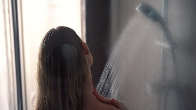 Young woman taking shower in modern glass shower cabin. Girl washing under running water in hotel or at home with big window. 