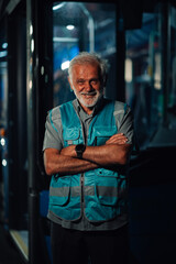 Senior bus driver smiling with crossed arms in depot at night