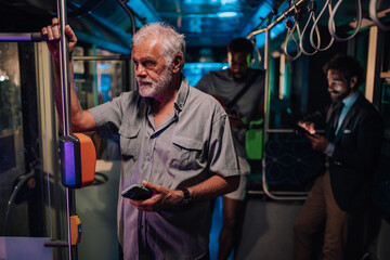 Senior man holding smartphone on a night bus with passengers around him