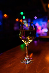 Glass of white wine on a wooden bar counter with colorful bokeh lights in the background. Cozy nightlife atmosphere in a modern nightclub or lounge bar