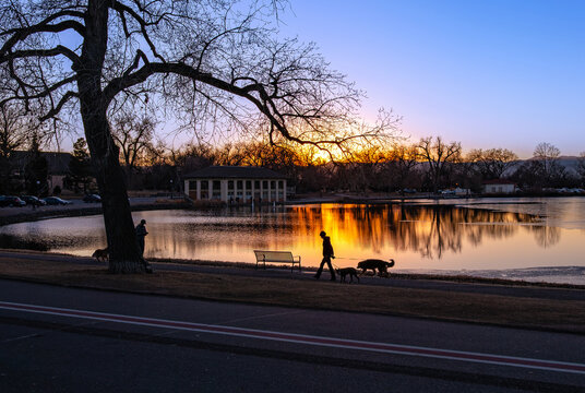 A backlit image of a dog walker at sunset with a colorful beam of golden light reflecting on the lake at Washington Park in Denver, Colorado.