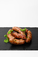A group of grilled homemade sausages garnished with fresh herbs, presented on a black slate plate. The golden-brown crust and visible meat texture highlight their hearty and appetizing look.