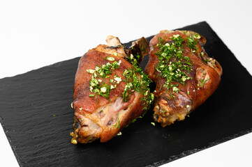 Two roasted pork knuckles with crispy brown crust, generously topped with fresh chopped dill and garlic, served on a stylish black slate plate over a clean white background