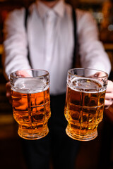 A waiter in a white shirt with suspenders holding two beer mugs filled with light beer. Bar or restaurant atmosphere with focus on drink and service