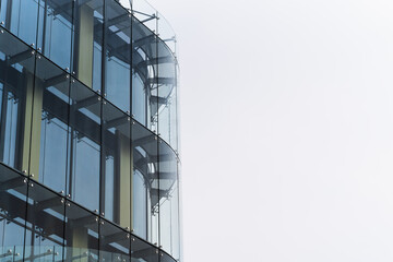 Photo shows the corner of a modern glass office building with reflective windows. The image...