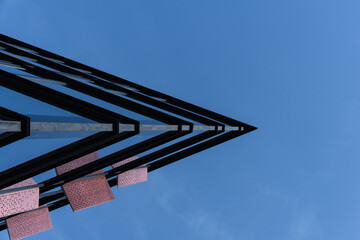Photo shows the sharp corner of a modern glass building with metal and pink panels, viewed from below. The image emphasizes architectural geometry and urban design against a clear blue sky.