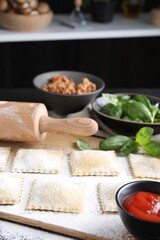 Uncooked ravioli, rolling pin, bolognese, sauce and basil on black table, closeup