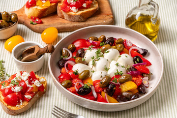 Caprese salad with colorful tomatoes, mozzarella balls, and a variety of olives, garnished with herbs, served in a bowl on a textured tablecloth with olive oil and bread