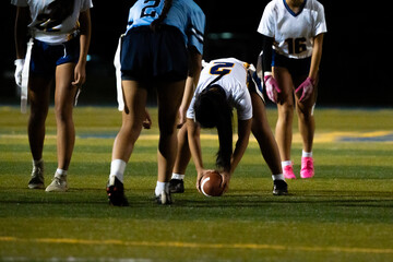 A young female center prepares to snap the football at the line of scrimmage during a nighttime flag football game. Focus, formation, and team readiness captured under stadium lights.