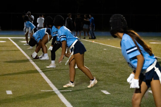 Girls flag football team lines up at scrimmage during nighttime game, players crouched and ready for the snap, showing focus, strategy, athletic discipline, and youth teamwork on turf.