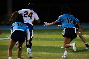 Girls flag football player with ball runs ahead as defenders in blue chase closely behind under night lights, intense youth athletic competition, teamwork, speed, and school spirit captured.