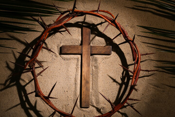 Wooden cross with crown of thorns and palm leaves on sand. Good Friday concept