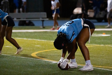 Girls flag football center prepares to snap the ball under lights, showing discipline, focus, and readiness in youth sports competition. Team positioning and intensity captured on turf at night.
