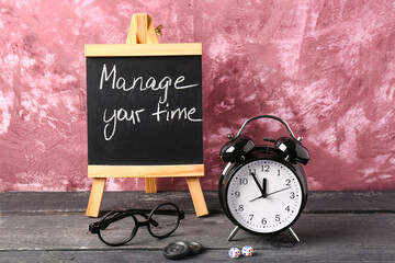 Blackboard with text MANAGE YOUR TIME,  alarm clock and glasses on wooden table against pink...
