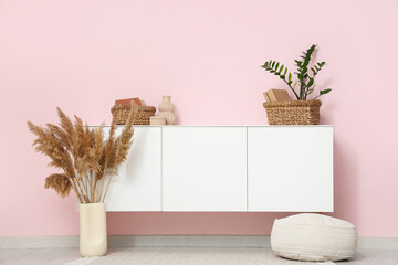 Chest of drawers, pampas grass, pouf and houseplant near pink wall in room