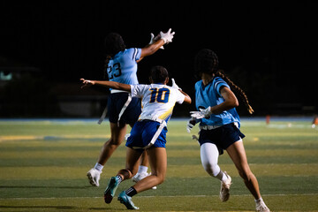 Girls flag football player leaps to catch the ball midair as defenders close in, high-intensity school sports moment under lights, showcasing youth athleticism, teamwork, focus, and determination.