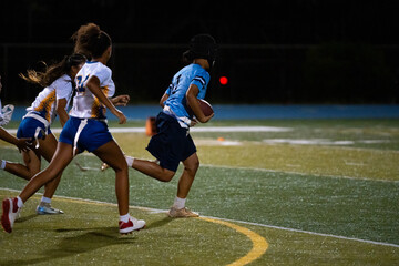 Girls flag football player runs toward end zone with ball as opponents chase in pursuit, intense youth sports game, school competition, team strategy, speed and agility under stadium lights.