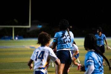 Girls flag football under stadium lights, player in blue jersey runs forward with ball while opponents chase, intense school game, teamwork, focus, youth athletics, night sports event captured.
