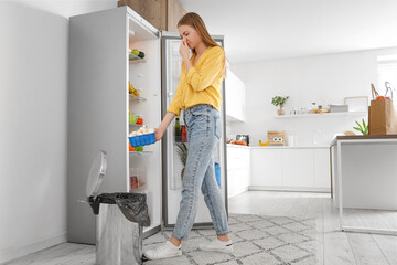 Young woman throwing spoiled mushrooms into trash bin near open fridge in kitchen