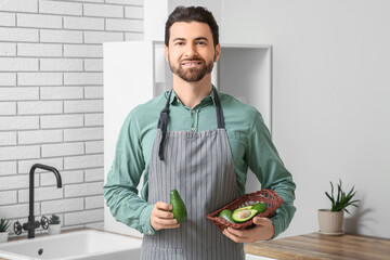 Handsome man holding basket with fresh avocado near sink in kitchen