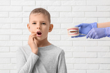Little boy with tooth ache and hands of dentist holding model of jaw on white brick background