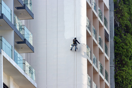 A professional worker with  safety equipment paints the tall exterior wall of a modern high-rise building - Powered by Adobe