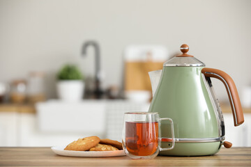 Modern green electric kettle with glass cup of hot tea and sweet cookies on wooden table in kitchen