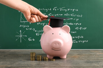 Woman putting graduation hat on piggy bank near green blackboard. Student loan concept