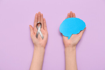 Woman holding grey awareness ribbon and paper brain on purple background. Cancer concept