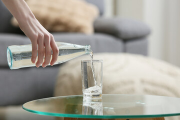 Woman pouring fresh water from bottle into glass on table at home, closeup