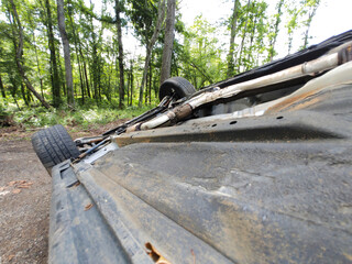 Rusty car rests on its side amidst trees foliage, significant damage from an accident.