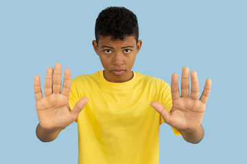Young Latino man with serious face wearing yellow shirt making stop sign with his hands on light blue background, teenager with a warning sign