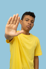 Vertical image of young Latino man wearing a yellow shirt with a serious face making a stop sign with his hand on a light blue background, teenager with a warning sign